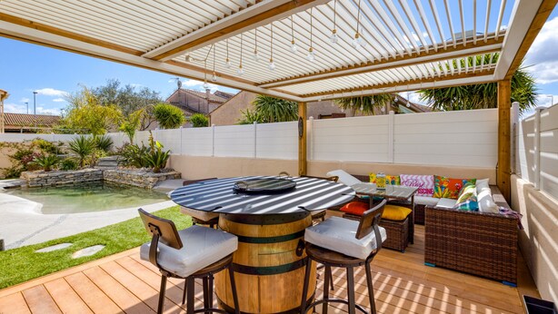 Een terras buiten met een houten tafel, barstoelen, een zachte zithoek en een zwembad.