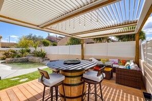 Une terrasse extérieure avec une table en bois, des tabourets de bar, un coin salon confortable et une piscine.