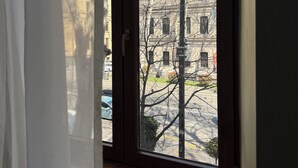 Blick durch ein Fenster mit Vorhang, auf eine Straße und ein Gebäude mit zahlreichen Fenstern.