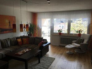 Una sala de estar con un sofá, una mesita de centro, una silla, una planta en maceta y una ventana con vistas a edificios.