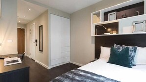 Een slaapkamer met een bed, een bureau, een kledingkast en een plank met decoratieve spullen.