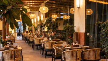Un comedor interior con sillas y mesas de mimbre, lámparas colgantes de mimbre y plantas en macetas.