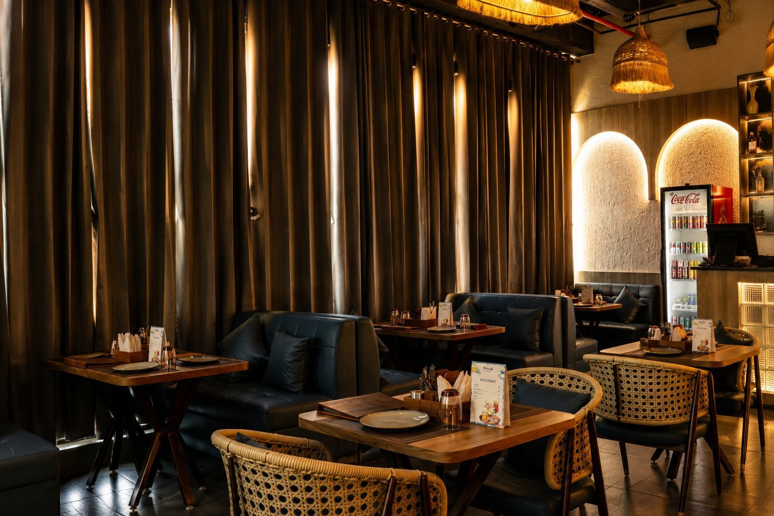 Un interior de restaurante con cortinas oscuras, asientos de cuero, sillas de mimbre y un refrigerador Coca-Cola.