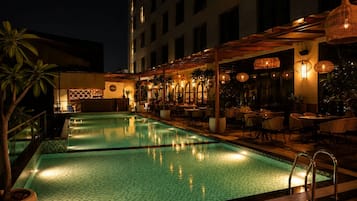Un área de piscina en la azotea con bar y asientos, rodeada de plantas en macetas e iluminación cálida.