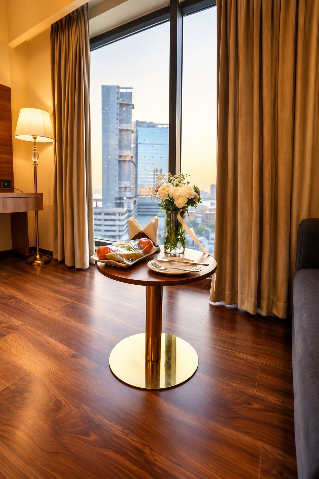 Une chambre d’hôtel avec une table à manger dressée pour deux personnes, un vase de fleurs et une vue sur la ville à travers de grandes fenêtres.