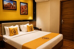 A hotel room with a bed, two pillows, a bedside table with a phone, and a door. - The Murti Hotel and Banquet (Greater Noida)