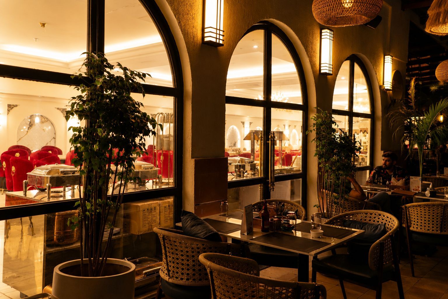 Un restaurante con amplias ventanas arqueadas, mobiliario de mimbre y plantas en macetas.