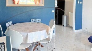 Uma área de refeições com mesa e cadeiras, uma janela com cortinas, um radiador e uma porta que dá para outro quarto.