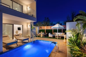 A modern outdoor pool area with lounge chairs, a small table, and a covered seating area. - Oasis Luxury Suites (Rhodes)