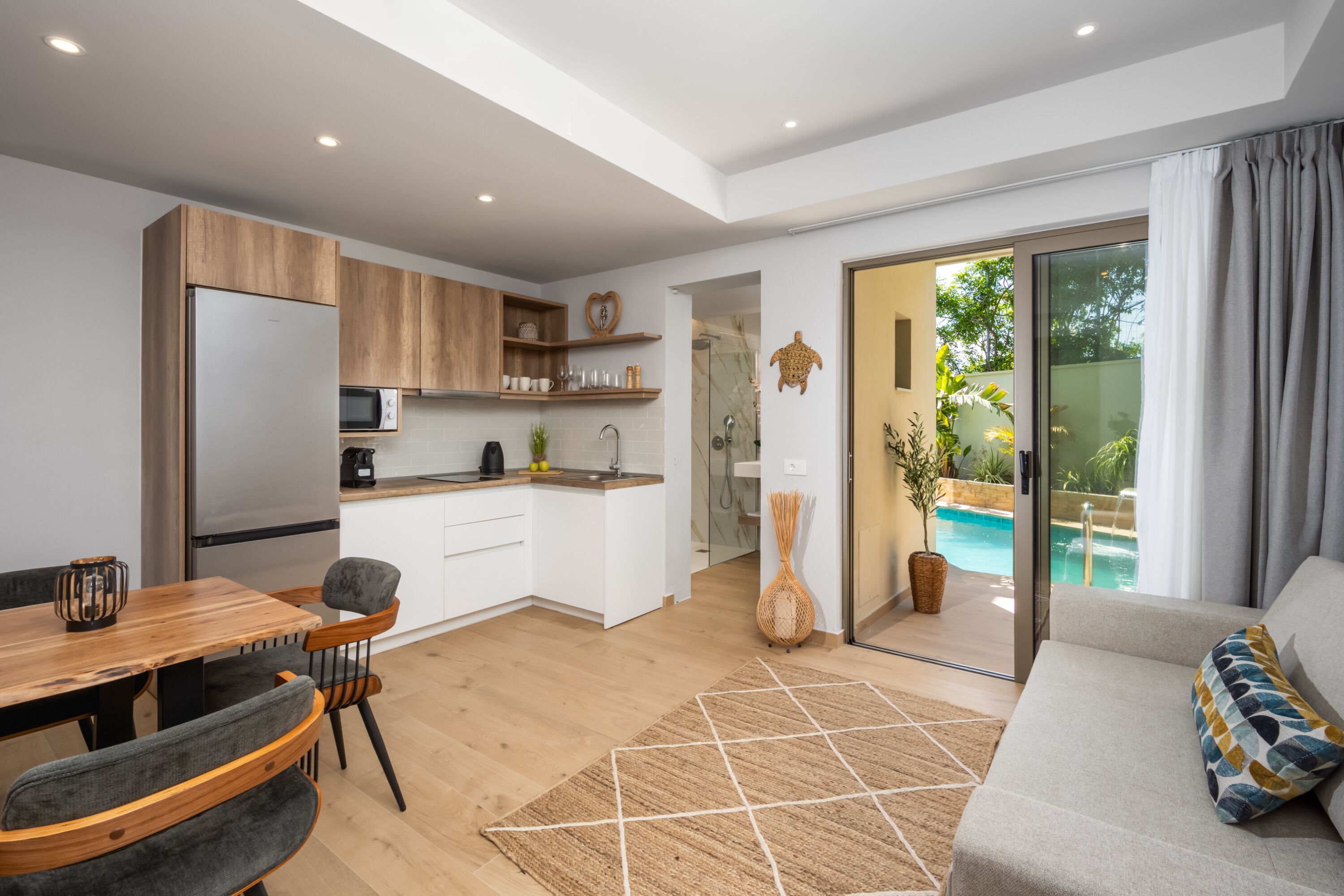 A modern kitchen with wooden cabinets, a dining area with a table and chairs, and a view of a swimming pool through a glass door.