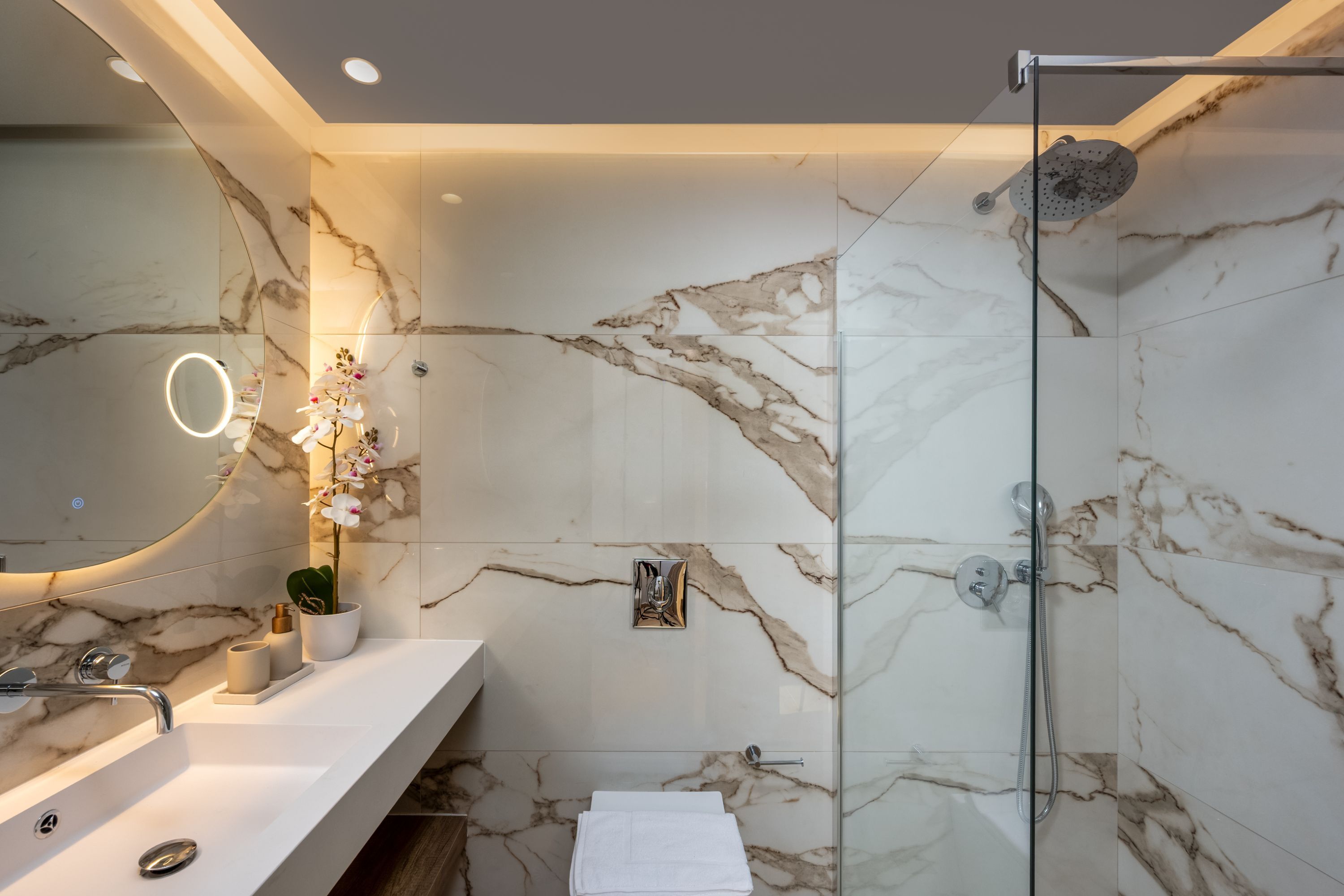 A modern bathroom with a glass shower enclosure, marble walls, a white sink, and a mirror with a ring light.