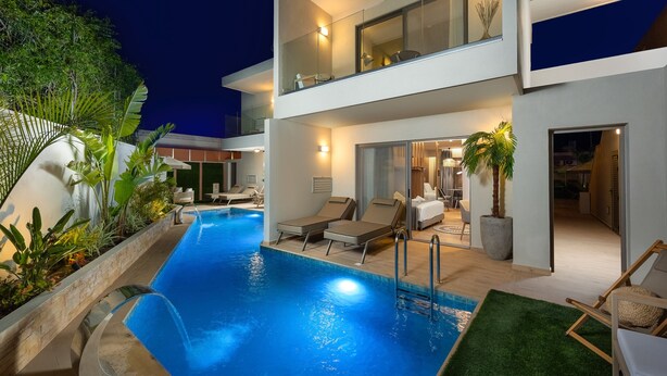 A modern two-story building with a swimming pool, lounge chairs, and a well-lit interior.