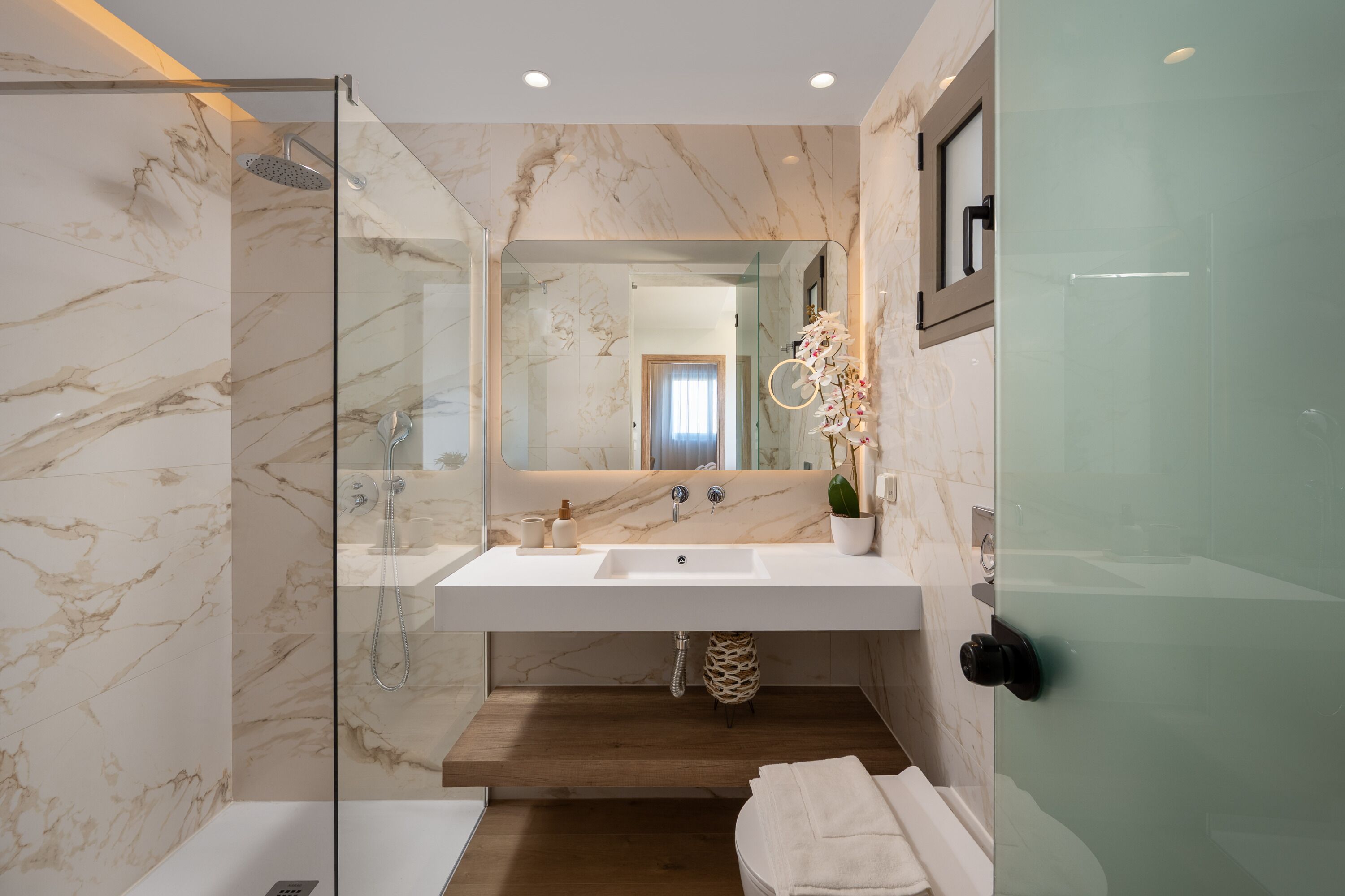 Ein modernes Badezimmer mit Duschkabine aus Glas, Marmorfliesen, einem weißen Waschbecken mit Spiegel und Holzboden.