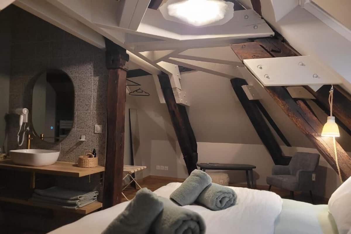 A bedroom with a bed, towels, a bedside table, a lamp, a chair, and a view of the outside.