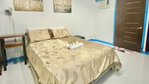 Un dormitorio con cama, mesita de noche, dos cuadros en la pared, una lámpara en el techo y una puerta.