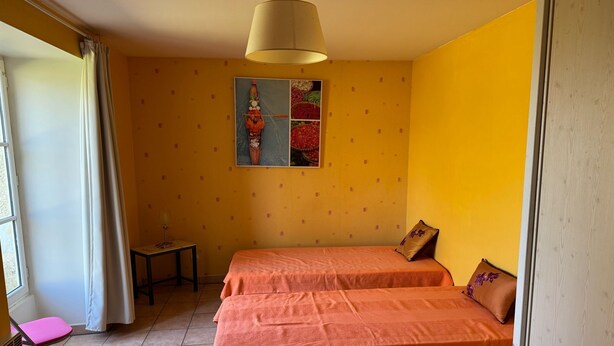 A room with two beds, a small table, a chair, a window with curtains, and a ceiling lamp. - Maison Indépendante (Vallon-sur-Gée)