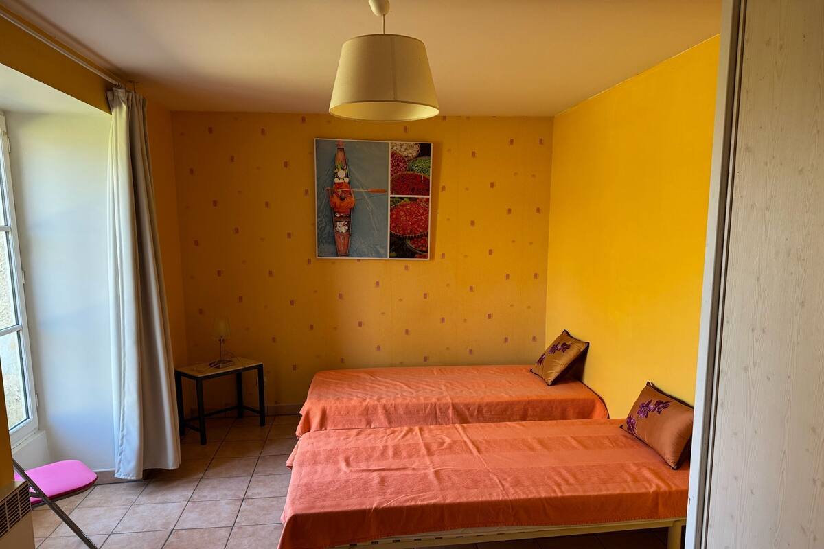 A room with two beds, a small table, a chair, a window with curtains, and a ceiling lamp.