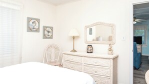 A bedroom with a bed, a dresser, a chair, a lamp, and wall art. - Register - A cozy 2-Bedroom Duplex with Soundfront Views for 6 Guests (Surf City)