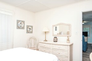 A bedroom with a bed, a dresser, a chair, a lamp, and wall art. - Register - A cozy 2-Bedroom Duplex with Soundfront Views for 6 Guests (Surf City)