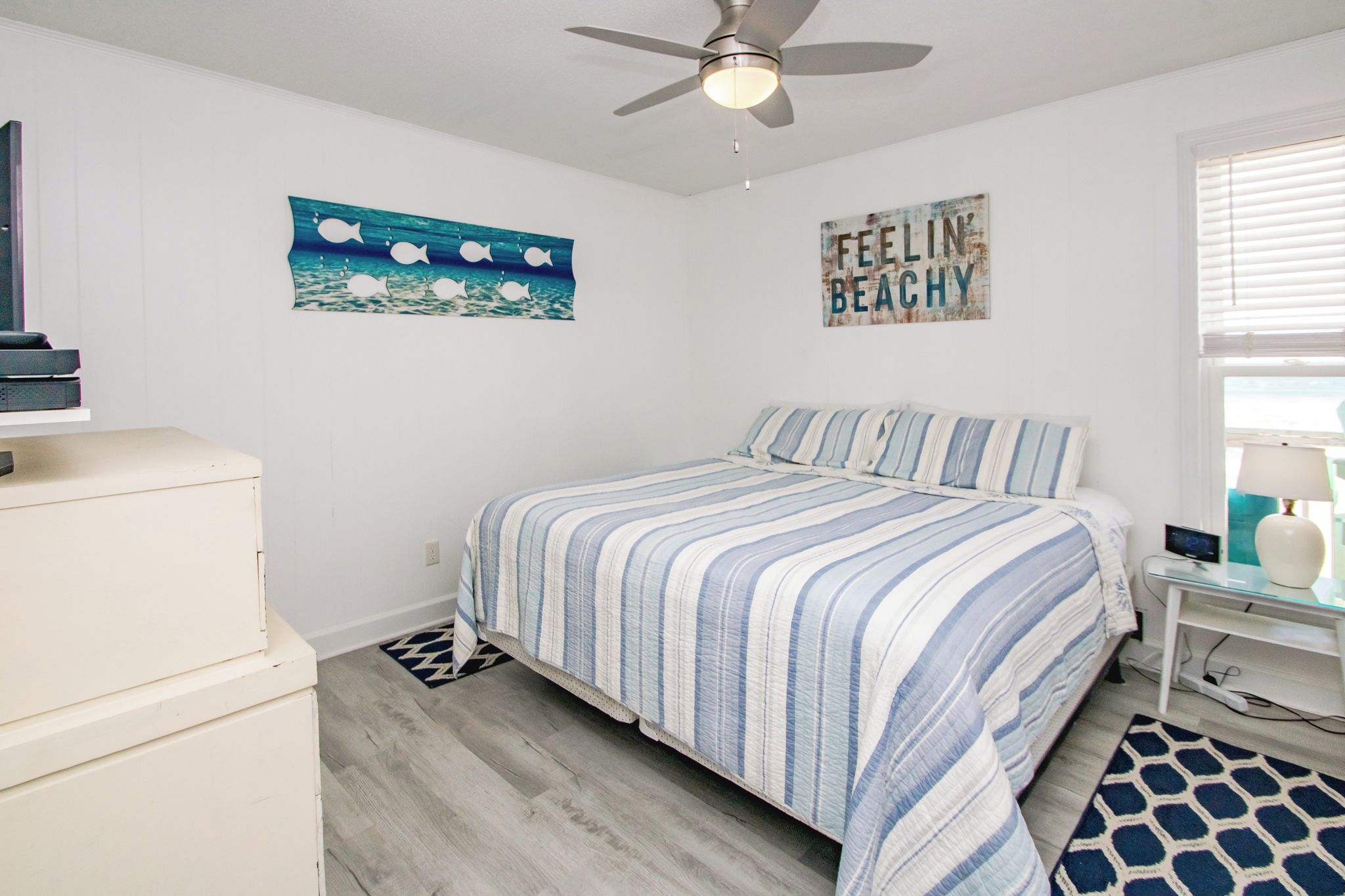 A bedroom with a bed, a ceiling fan, a wall with a 'FEELIN' sign, and a window with blinds.