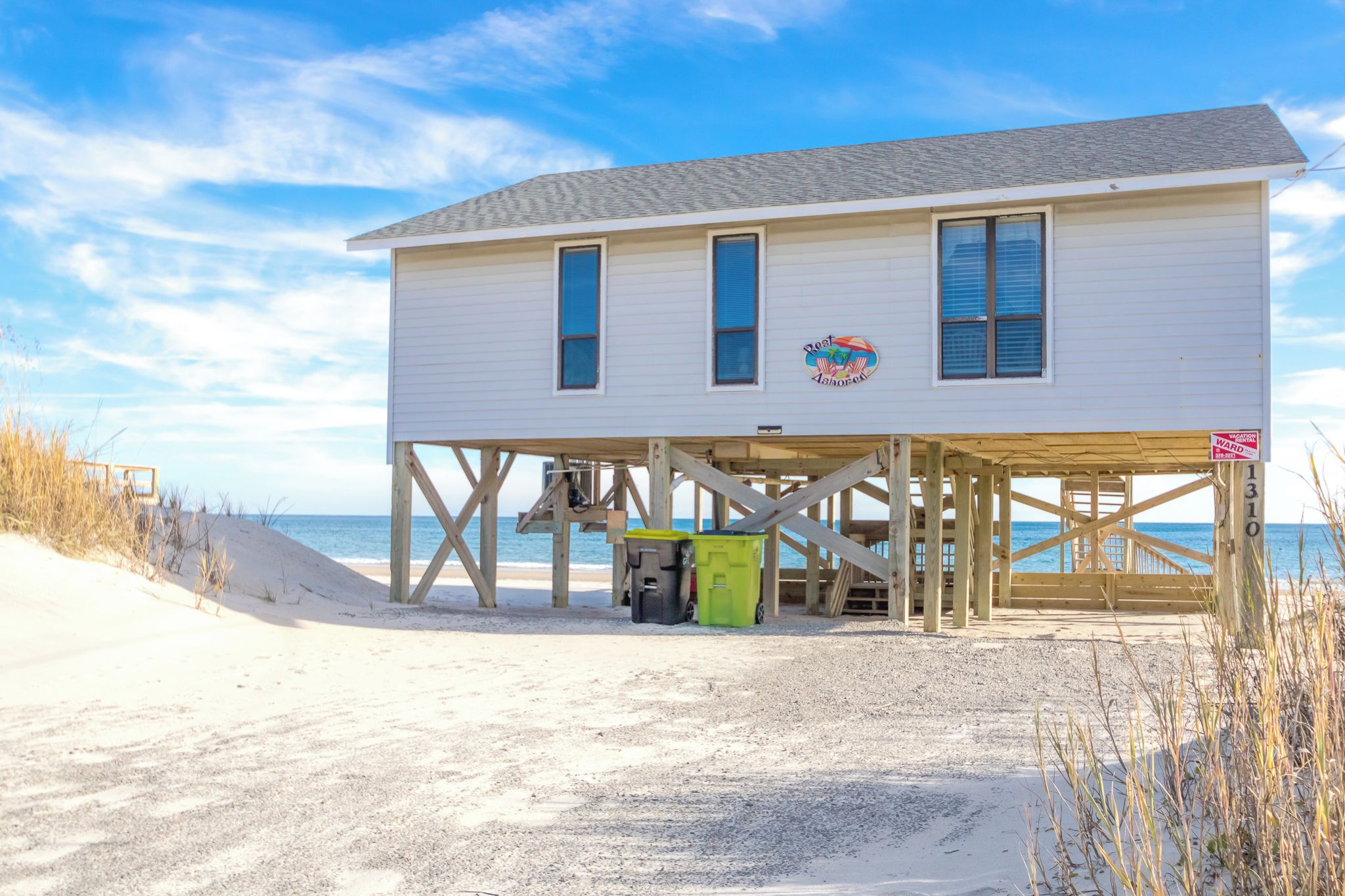 A beach house with a wooden deck, a beach view, and a sign indicating vacation rental availability.