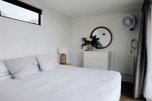 Une chambre à coucher comprenant un lit, une table de chevet avec une lampe, une commode, un miroir et un ventilateur fixé au mur.
