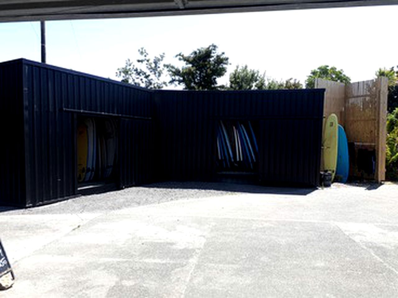 A covered outdoor area with a concrete floor, a dark blue structure, and a surfboard leaning against the wall.