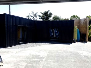 Un espacio al aire libre techado con piso de concreto, una estructura azul oscuro y una tabla de surf apoyada en la pared.