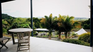 An outdoor patio with a round table and chairs, surrounded by lush greenery and a view of distant hills. - Luxury Camping Pod for Two to Go Glamping near Auckland (Te Arai)