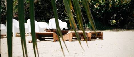 Eine Strandlounge mit weißen Plastikstühlen und Holzliegen, umgeben von üppiger Bepflanzung.