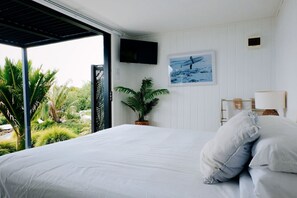 Une chambre à coucher avec un lit, un téléviseur fixé au mur, une plante en pot et une vue sur la verdure à l’extérieur.