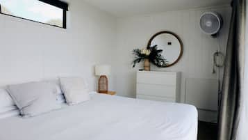 A bedroom with a bed, a bedside table with a lamp, a dresser, a mirror, and a wall-mounted fan.