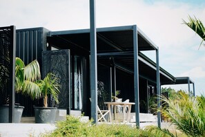 Eine moderne Außenarchitektur mit überdachtem Sitzbereich, großen Pflanzgefäßen mit tropischen Pflanzen und blauem Himmel.