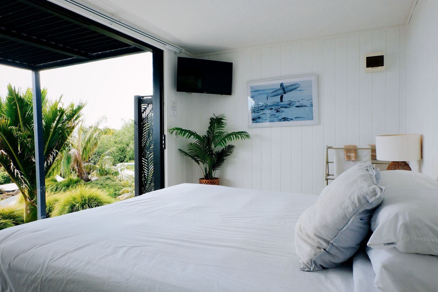 A bedroom with a bed, a TV mounted on the wall, a potted plant, and a view of greenery outside.