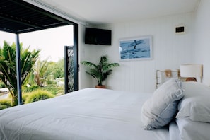A bedroom with a bed, a TV mounted on the wall, a potted plant, and a view of greenery outside.