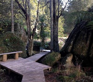 A wooden pathway in a forested area with benches and a large rock formation. - Luxury Accommodation near Whangarei Ideal for a North Island Weekend Getaway (Ruatangata West)