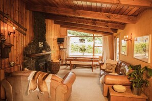A cozy cabin interior with a stone fireplace, wooden walls, a dining area, and a living room furnished with leather sofas and a coffee table.