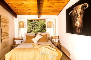 A bedroom with a bed, pillows, a blanket, a wooden ceiling, a window with a view of trees, and a wall-mounted sculpture.