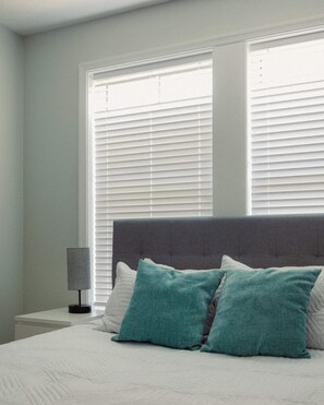 A bedroom with a bed, two windows with blinds, a bedside lamp, and a grey headboard. - Villa Mojito Apartment A in Historic Downtown (St. Augustine)