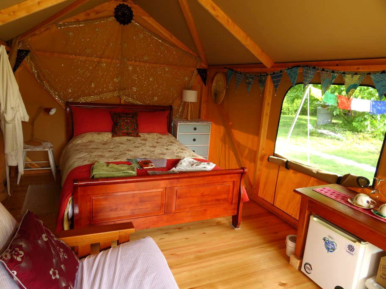 A tent-style bedroom with a wooden bed, a small desk, and a window with a view of greenery.