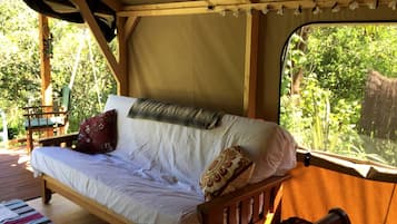 Un lit en bois avec une literie et des oreillers blancs, une fenêtre donnant sur un paysage verdoyant et une terrasse en bois à l’extérieur.