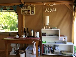 A cozy kitchen area with a wooden table, a kettle, a toaster, and a fan. There is a bookshelf with books and a sign that reads 'ALOHA'. - Glamping Tent on Permaculture Farm near Pohara Beach in Golden Bay, South Island (Pohara)