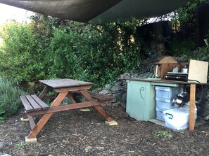 Una mesa de picnic y una pequeña área de cocina al aire libre, con un muro de piedra y follaje en el fondo.
