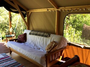 A wooden bed with white bedding and pillows, a window with a view of greenery, and a wooden deck outside.