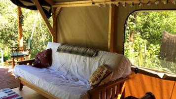 A wooden bed with white bedding and pillows, a window with a view of greenery, and a wooden deck outside.