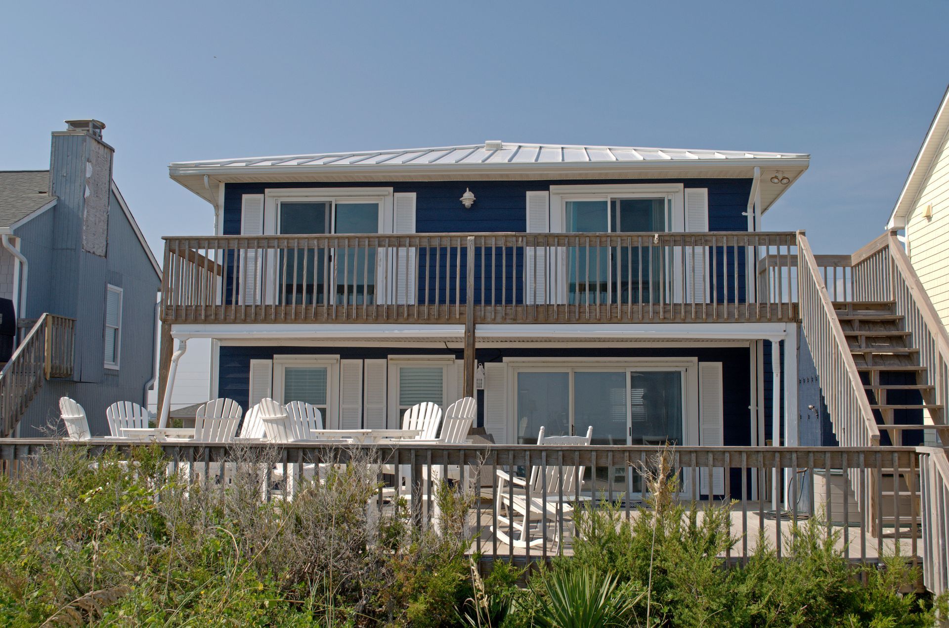 Une maison à deux étages située en bord de mer, dotée d’un balcon, de meubles d’extérieur blancs et d’un escalier en bois.
