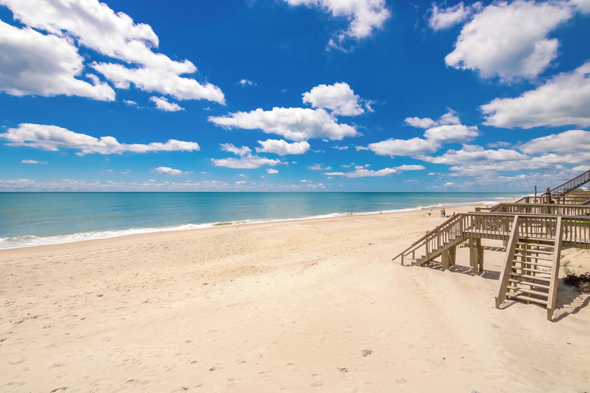 Une plage avec un escalier en bois menant à une structure également en bois, d’où l’on peut admirer une mer bleue et limpide.
