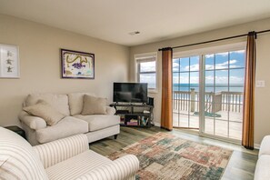 A living room with a sofa, a television, and a view of the beach through large windows. - Sea-Renity Oceanfront 4 bedroom, 2 bath home in Surf City! (Surf City)