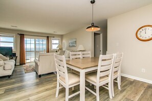 Un coin repas avec une table en bois et des chaises blanches, une horloge au mur et une vue sur l’océan par de grandes fenêtres.