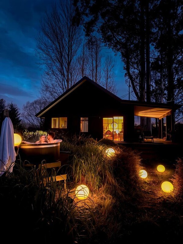 A modern house with a hot tub and outdoor seating area, surrounded by tall trees and illuminated by spherical lights.